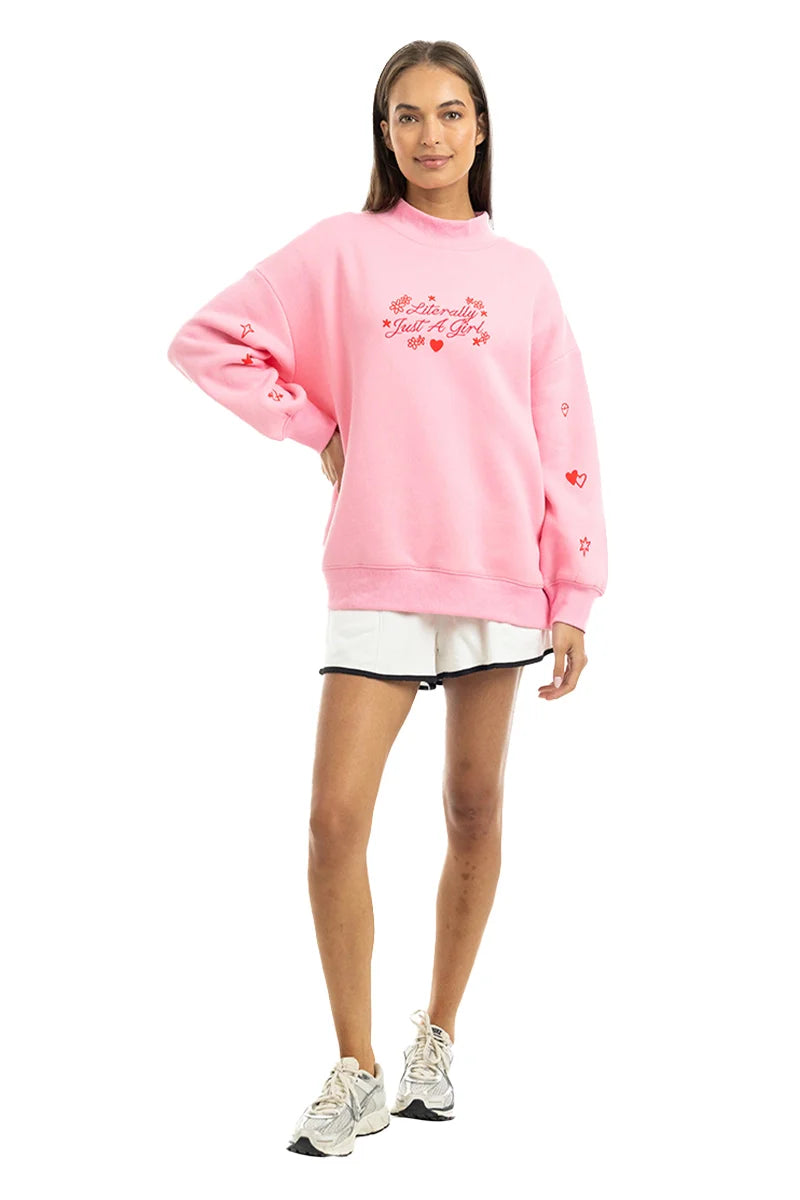 A woman with long brown hair smiles in the Katydid Literally Just A Girl Mockneck Pullover, a cute pink oversized sweatshirt with red embroidery, styled with white shorts and sneakers against a plain white background.