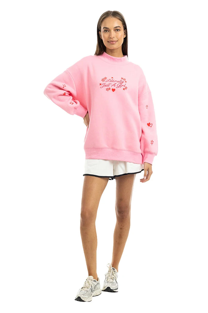 A woman with long brown hair smiles in the Katydid Literally Just A Girl Mockneck Pullover, a cute pink oversized sweatshirt with red embroidery, styled with white shorts and sneakers against a plain white background.