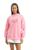 A woman smiles in the Katydid Literally Just A Girl Mockneck Pullover, a cute pink embroidered sweatshirt with red graphics and text on the front, paired with white shorts against a plain white background.