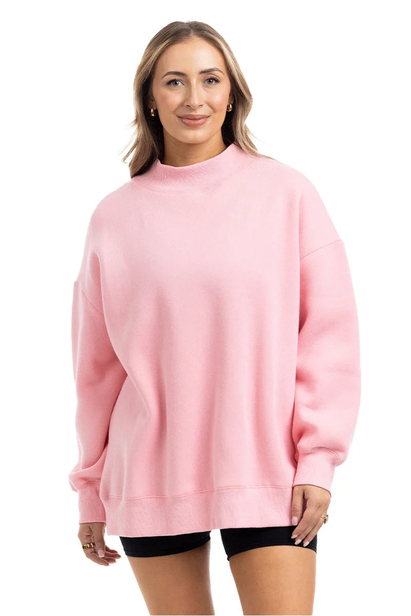 A woman faces forward, smiling, wearing the Katydid Pink Essential Mockneck Crew with black shorts against a plain white background.