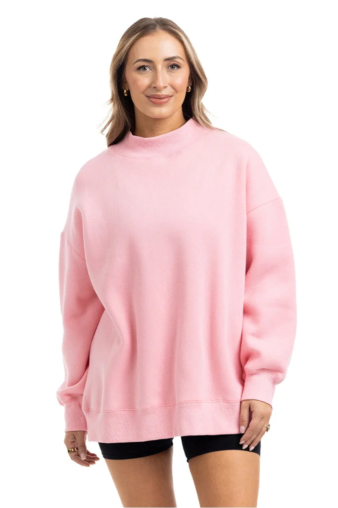 A woman faces forward, smiling, wearing the Katydid Pink Essential Mockneck Crew with black shorts against a plain white background.
