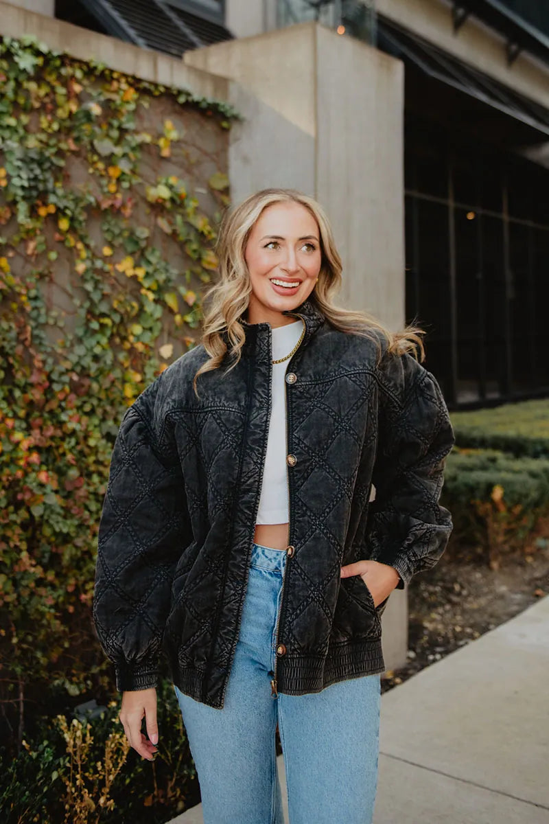 A woman with long, wavy blonde hair smiles by an ivy-covered wall, wearing the Katydid Womens Charcoal Luxe Oversized Jacket over a white crop top and blue jeans, with one hand in her jacket pocket.