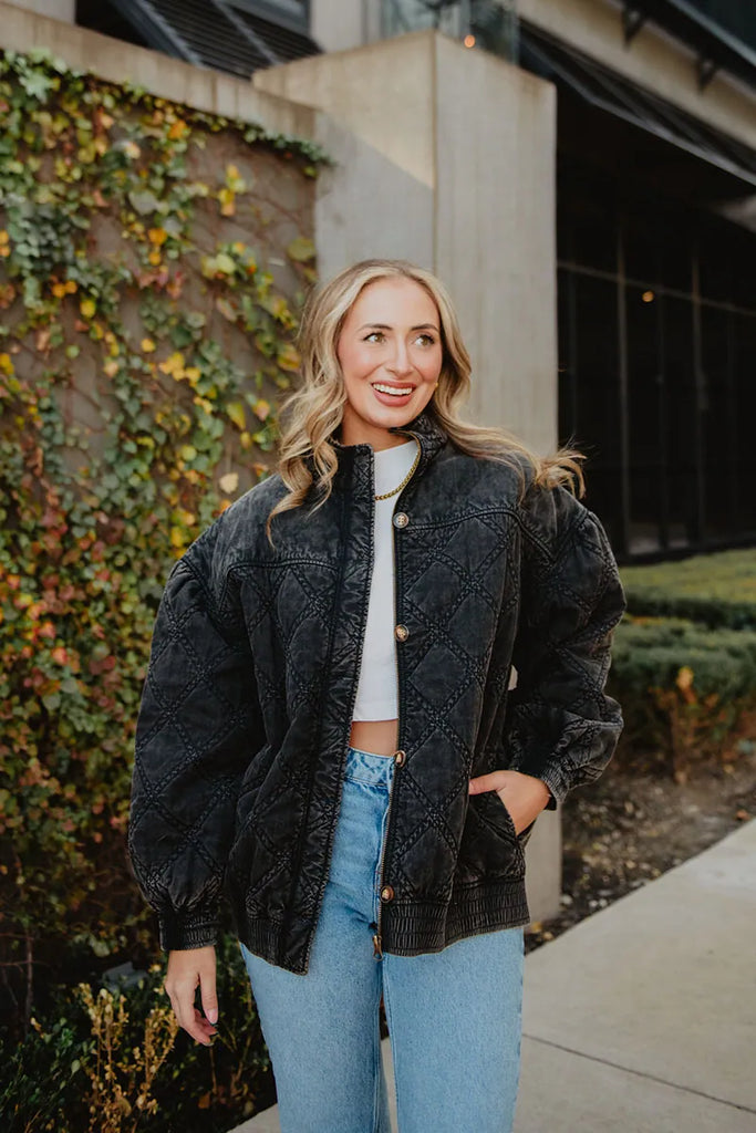 A woman with long, wavy blonde hair smiles by an ivy-covered wall, wearing the Katydid Womens Charcoal Luxe Oversized Jacket over a white crop top and blue jeans, with one hand in her jacket pocket.