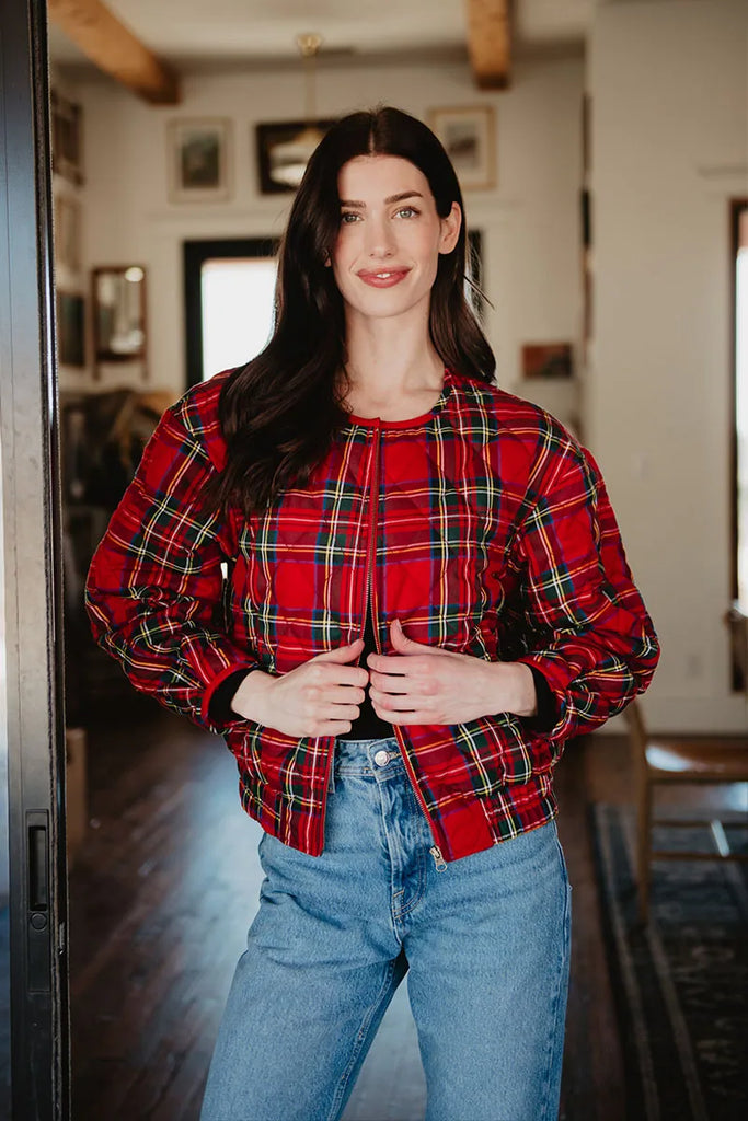 A woman with long dark hair smiles indoors, wearing the Katydid Royal Stewart Plaid Holiday Quilted Jacket over a black top and blue jeans, in a room with wooden beams, framed art, and gentle natural light.