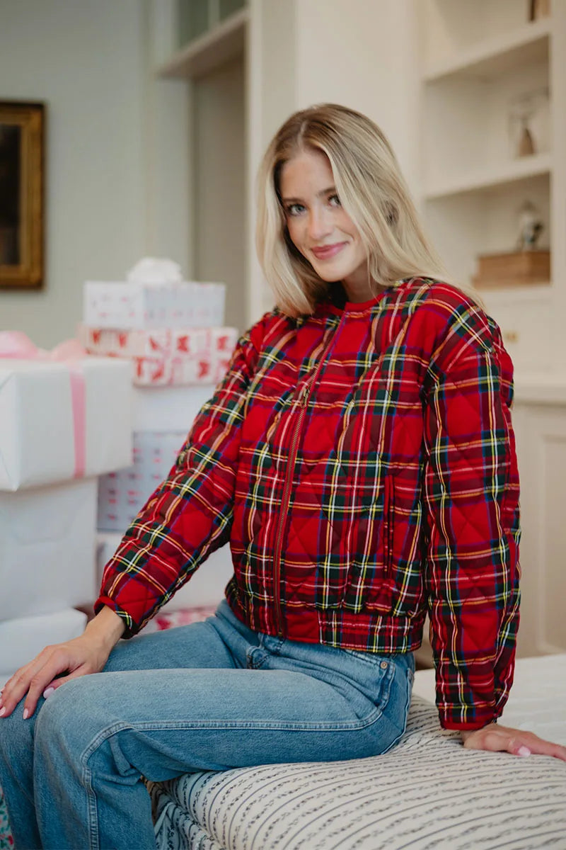 A woman with long blonde hair, wearing the Katydid Royal Stewart Plaid Holiday Quilted Jacket and blue jeans, sits on a bed with festive wrapped gifts behind her, creating a cheerful holiday atmosphere.
