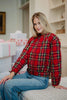 A woman with long blonde hair, wearing the Katydid Royal Stewart Plaid Holiday Quilted Jacket and blue jeans, sits on a bed with festive wrapped gifts behind her, creating a cheerful holiday atmosphere.