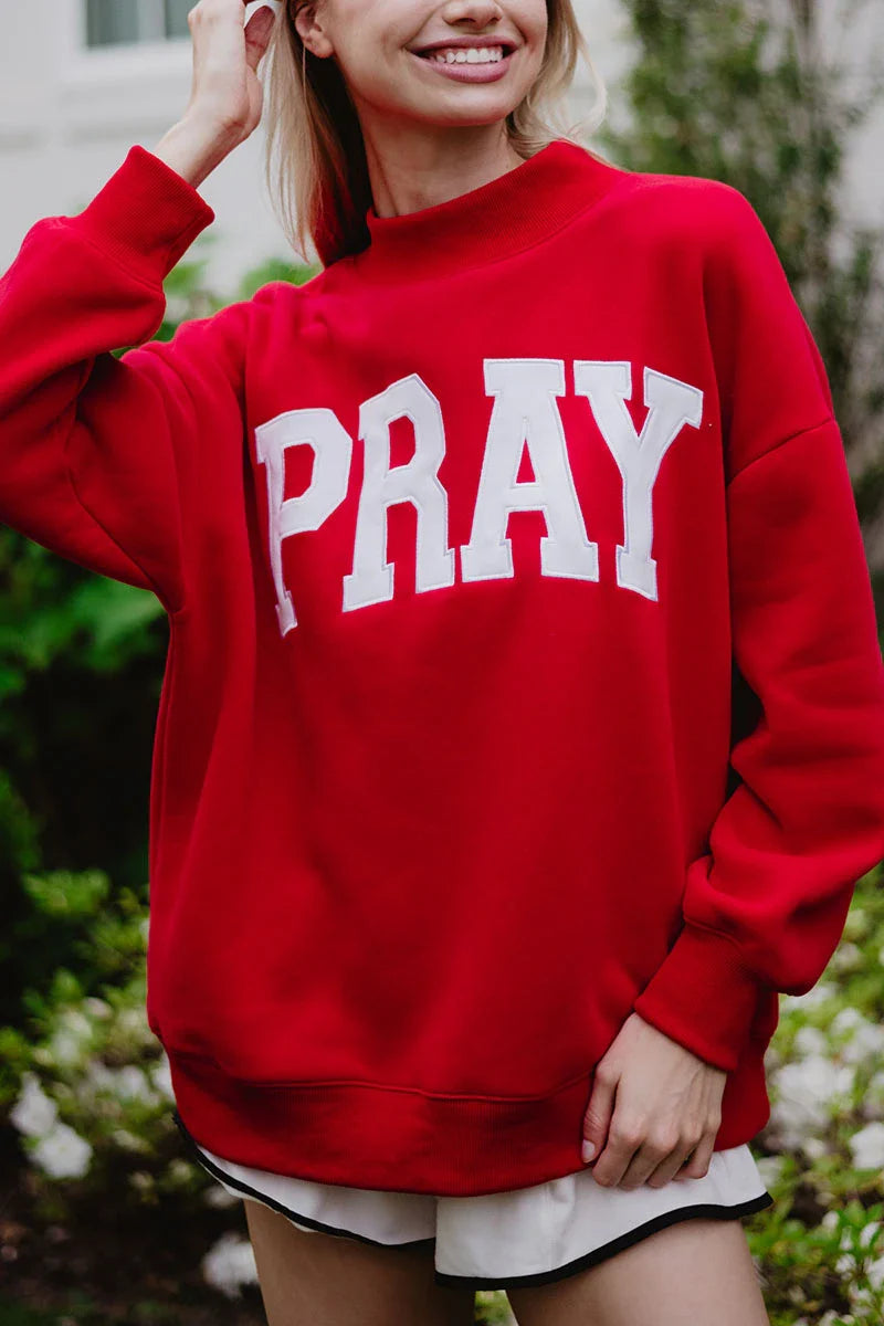 A smiling person wears the Katydid PRAY Mockneck Sweatshirt, featuring bold white PRAY lettering, while standing outdoors amid greenery.