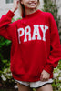 A smiling person wears the Katydid PRAY Mockneck Sweatshirt, featuring bold white PRAY lettering, while standing outdoors amid greenery.