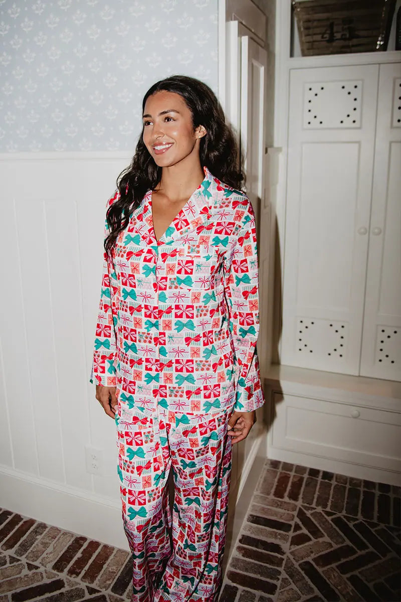 A woman smiles indoors wearing the Katydid Wrapped Presents with Bows Pajama Pants Set. She stands in a white-paneled room with a brick floor and built-in cabinets in the background.