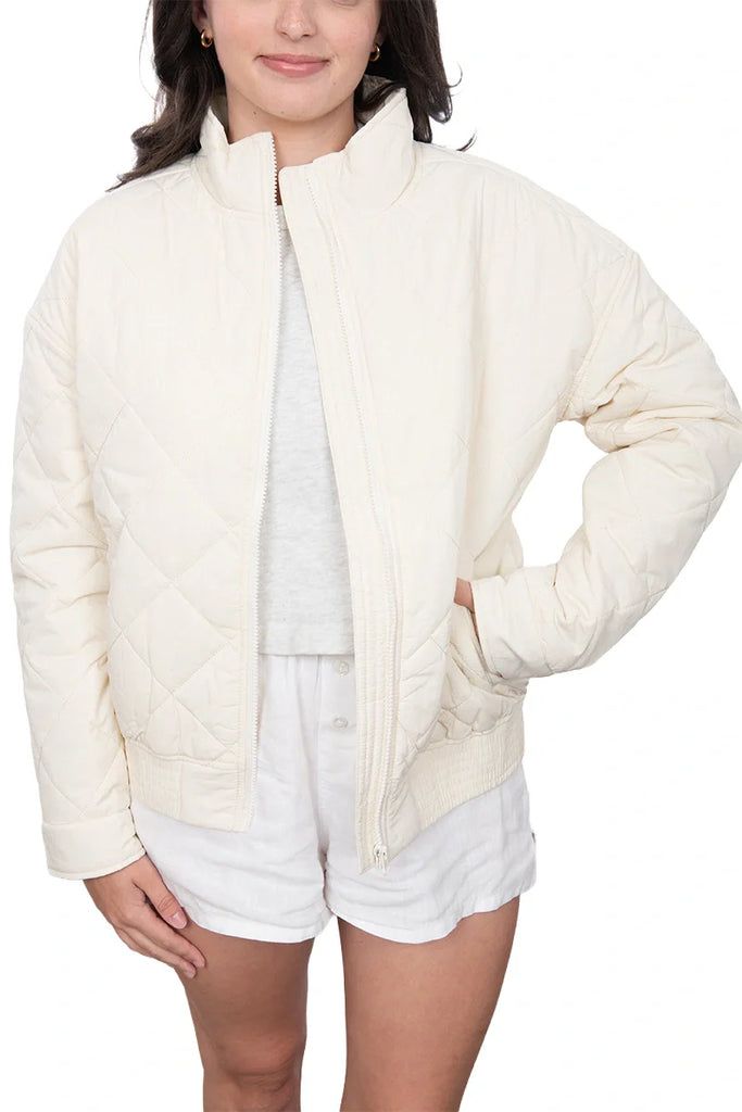 A person wearing the Katydid Cloud White Sporty Diamond Quilted Jacket over a white t-shirt and shorts stands with one hand in their pocket against a plain background.