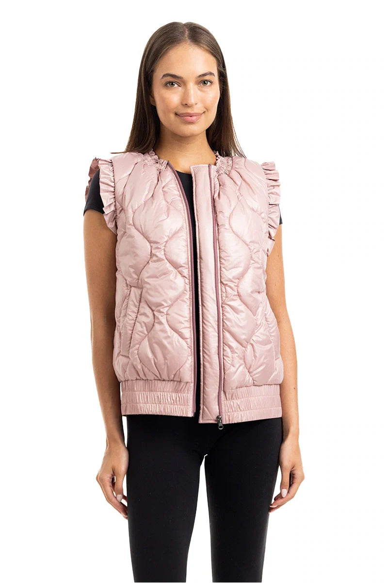 A woman with straight brown hair wears the Katydid Blush Metallic Puffy Vest with ruffle shoulder details over a black top and pants, standing against a white background.