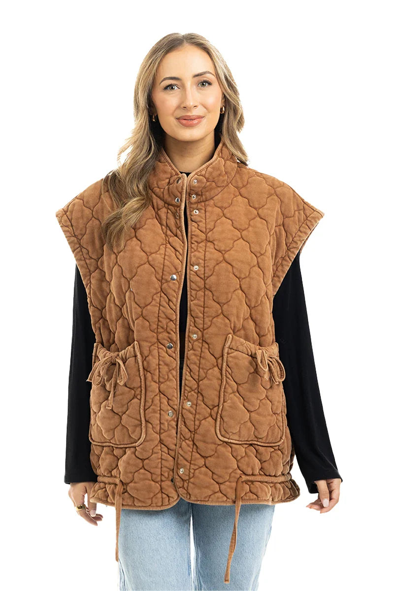 A woman models the Katydid Brown Oversized Womens Quilted Vest, styled with a black long-sleeve shirt and light blue jeans, smiling as she stands against a white background.