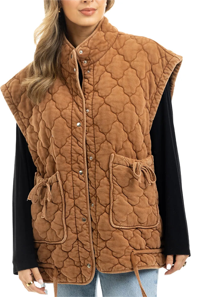 A woman styles the Katydid Brown Oversized Women’s Quilted Vest, featuring snap buttons, large front pockets, a high collar, and a padded pattern, over a black long-sleeve shirt and light blue jeans.