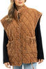 A woman styles the Katydid Brown Oversized Women’s Quilted Vest, featuring snap buttons, large front pockets, a high collar, and a padded pattern, over a black long-sleeve shirt and light blue jeans.