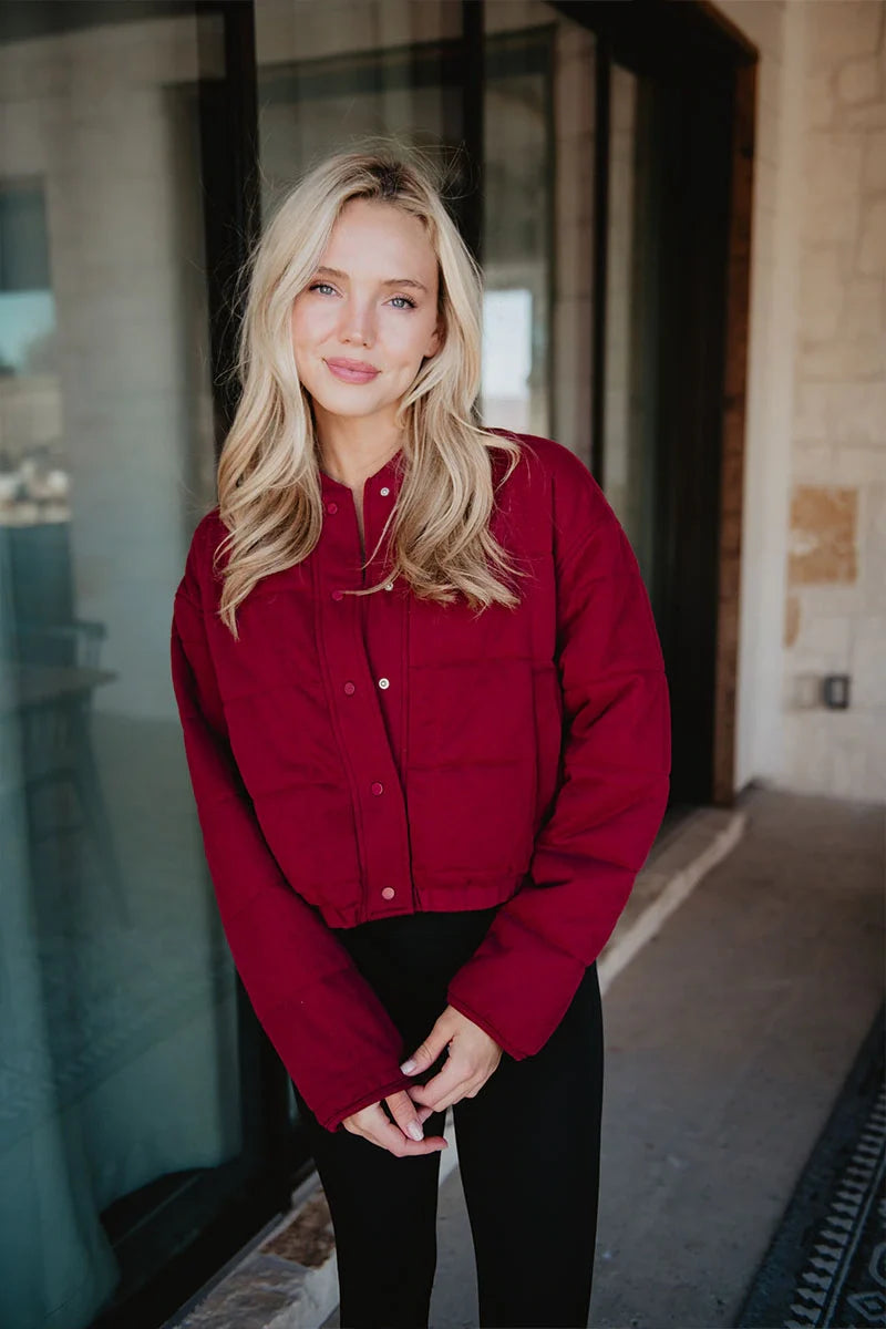 A woman with long blonde hair smiles softly at the camera outside a building, wearing the Katydid Womens Wine Cropped Quilted Bomber Jacket and black pants.