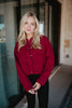 A woman with long blonde hair smiles softly at the camera outside a building, wearing the Katydid Womens Wine Cropped Quilted Bomber Jacket and black pants.