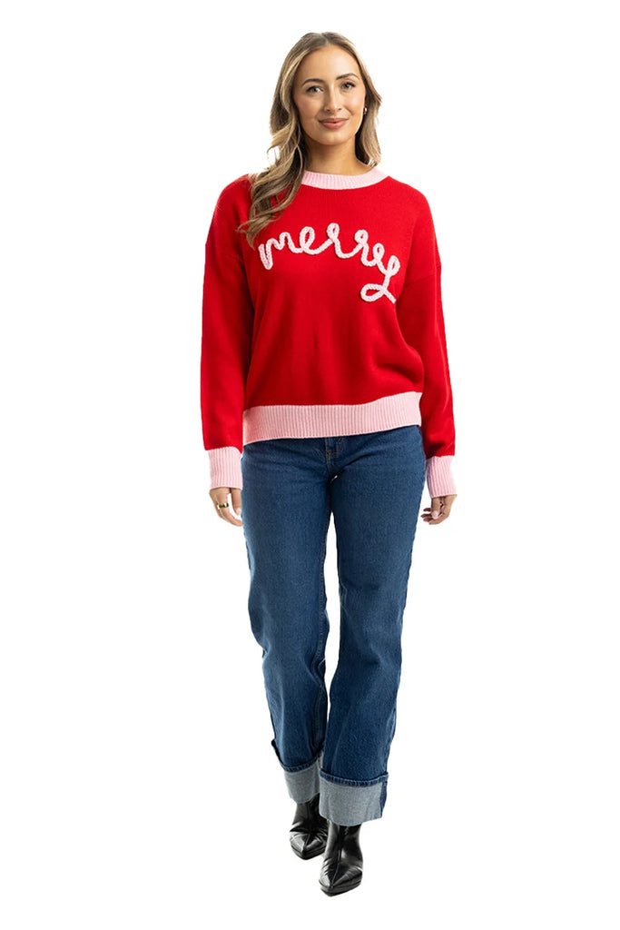 A woman smiles in the Katydid Merry Embroidered Sweater Christmas Woman, styled with blue cuffed jeans and black boots, posing against a white background.