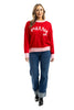 A woman smiles in the Katydid Merry Embroidered Sweater Christmas Woman, styled with blue cuffed jeans and black boots, posing against a white background.