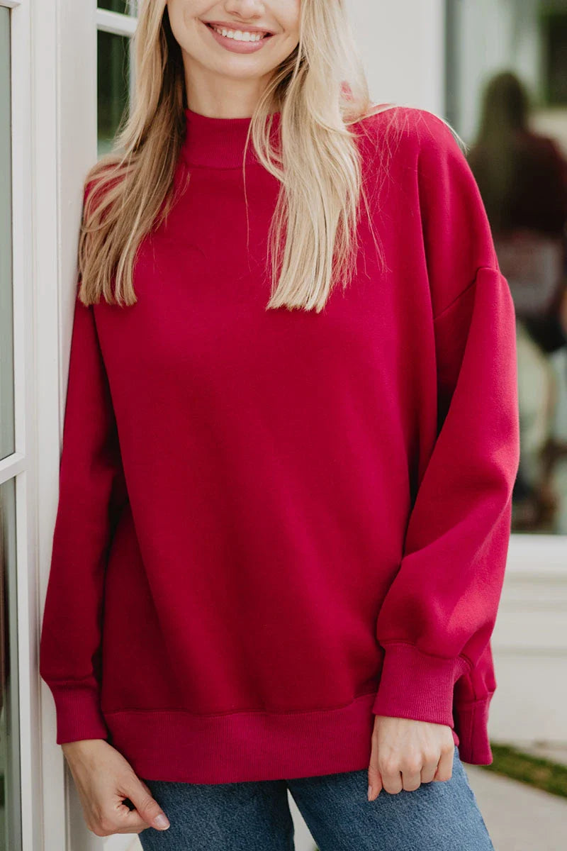 A woman with long blonde hair, smiling near a white door, wears the Katydid Red Essential Solid Mockneck with blue jeans. The softly blurred background enhances the cozy fall wardrobe vibe.