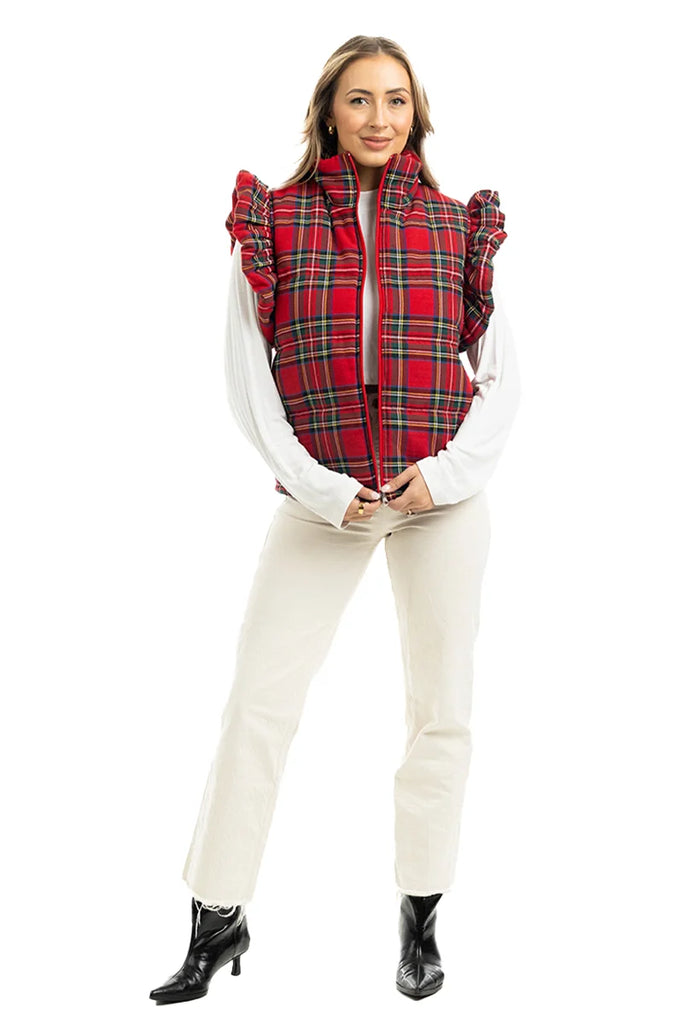 A person faces forward in a Katydid Royal Stewart Plaid Ruffle Puffer Vest over a white long-sleeve top, cream pants, and black ankle boots, creating a festive holiday look against a plain white background.
