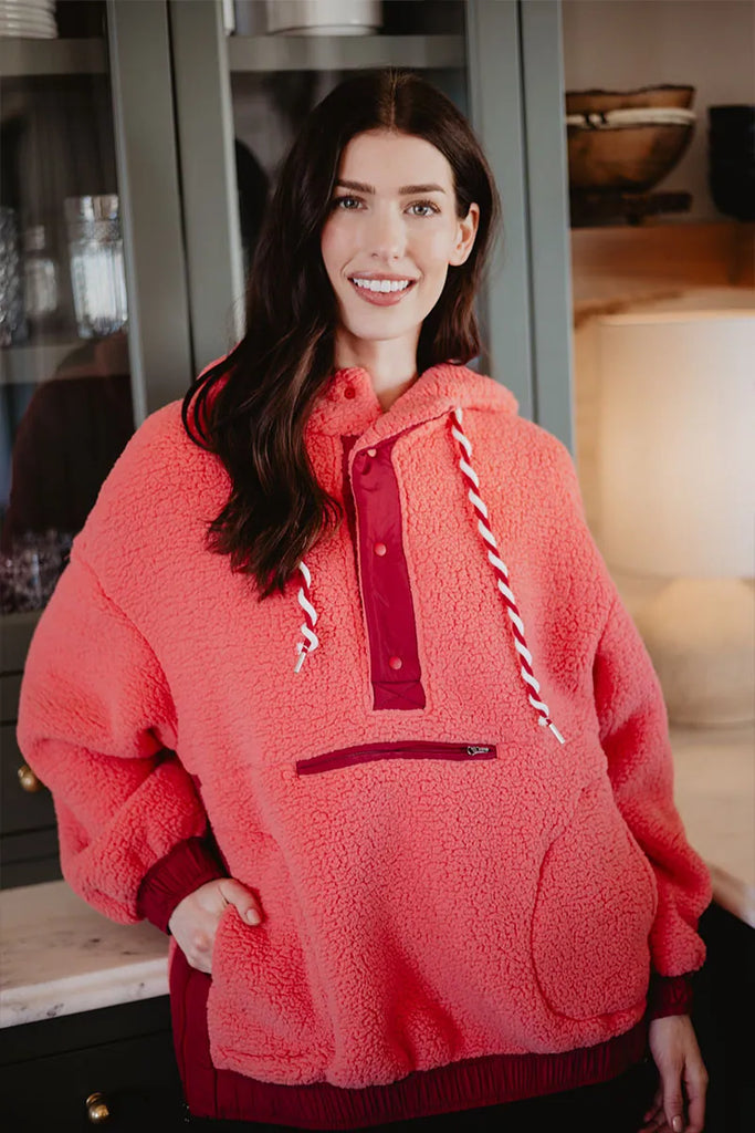 A woman with long dark hair smiles indoors near dark cabinets and a marble countertop, wearing the Katydid Coral Two-Toned Womens Fleece Pullover.