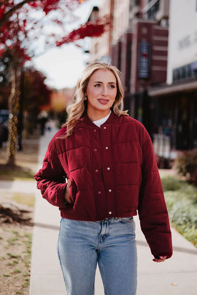 A blonde woman walks on a city sidewalk lined with trees and buildings, wearing blue jeans and the Katydid Womens Wine Cropped Quilted Bomber Jacket.