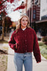 A blonde woman walks on a city sidewalk lined with trees and buildings, wearing blue jeans and the Katydid Womens Wine Cropped Quilted Bomber Jacket.