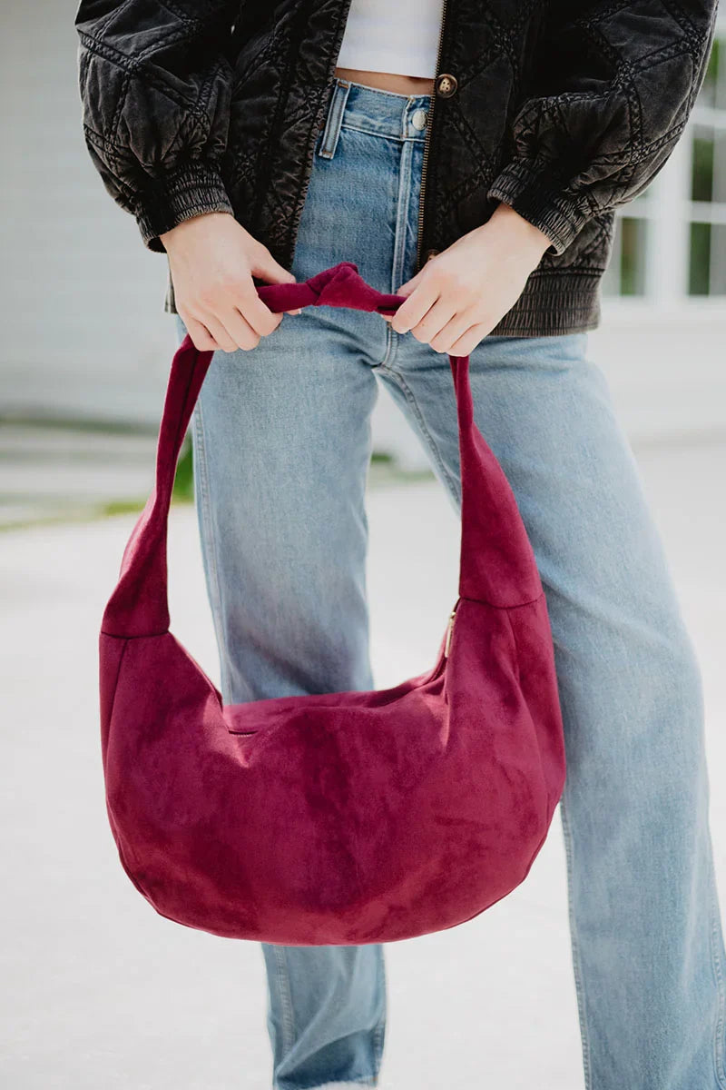 A person in a black jacket and blue jeans holds the Katydid Wine Casual Suede Tote, featuring a soft shape, thick strap, and deep red suede—a perfect fall accessory for any outfit.