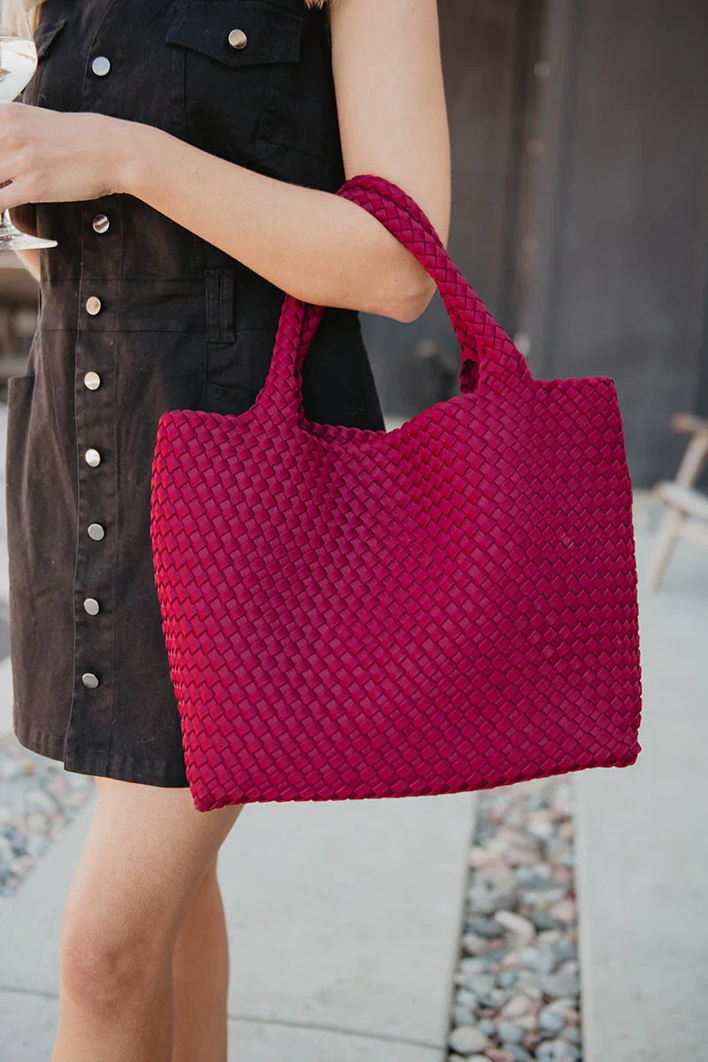 A woman in a sleeveless black button-up dress holds the Katydid Wine Vibrant Woven Tote Bag while standing outdoors on a sidewalk near rocks, with part of a building visible in the background.