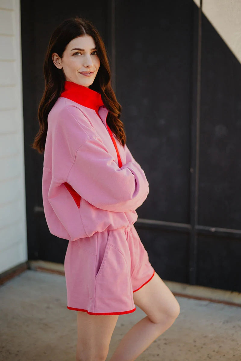 A woman with long brown hair stands outdoors, smiling and posing sideways with arms crossed in front of a dark wall while wearing the Katydid Pink  Orange Quarter Snap 2 Piece Athletic Set made from ultra-soft performance fabric.