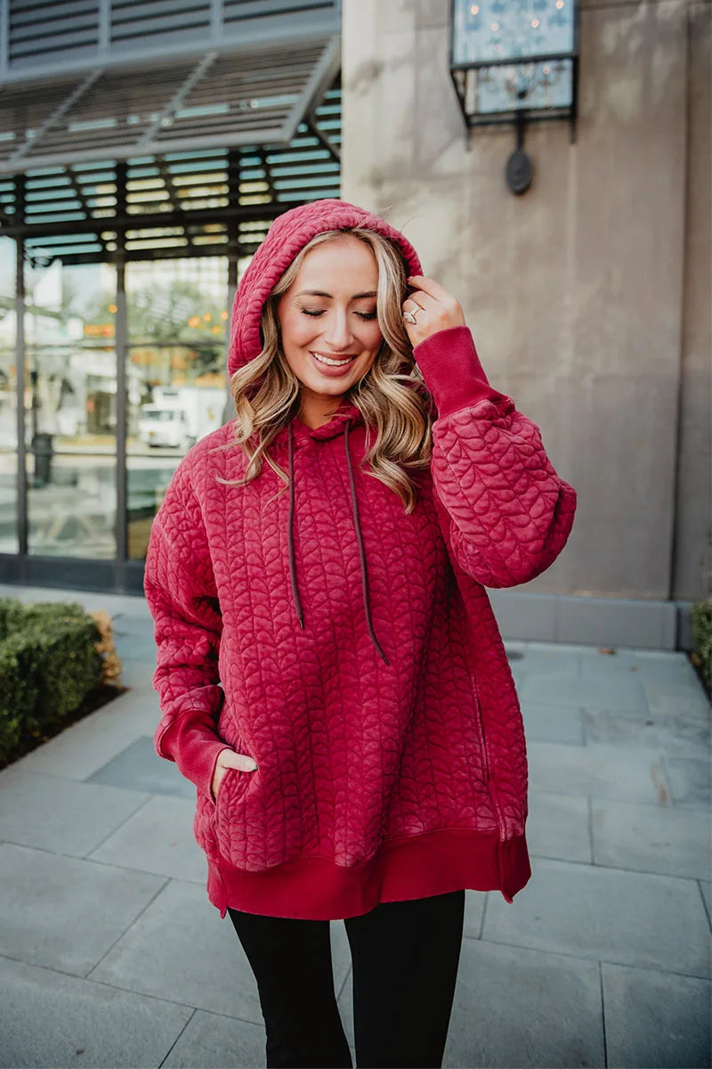 A woman enjoys a cozy, relaxed moment outdoors in the Katydid Red Heart Quilted Garment Washed Hoodie for Women, hood up and one hand in her pocket, smiling with her eyes closed and wearing black pants.