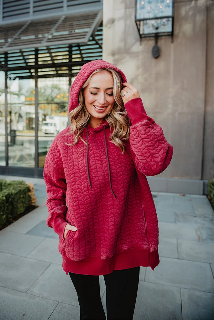 A woman enjoys a cozy, relaxed moment outdoors in the Katydid Red Heart Quilted Garment Washed Hoodie for Women, hood up and one hand in her pocket, smiling with her eyes closed and wearing black pants.