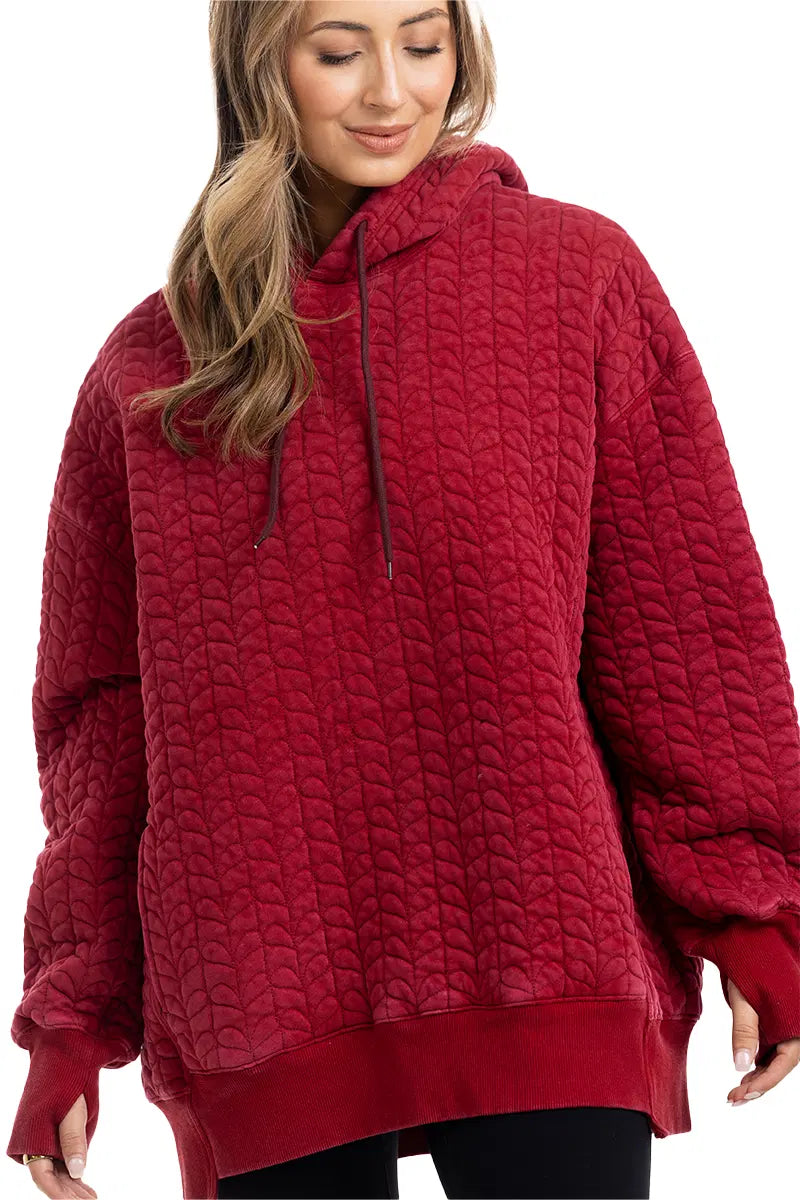 A woman models the Katydid Red Heart Quilted Garment Washed Hoodie, an oversized, casual piece, standing against a white background and looking down with a slight smile.