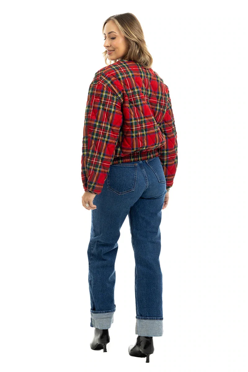 A woman stands smiling with her back to the camera, wearing Katydids Royal Stewart Plaid Holiday Quilted Jacket, blue cuffed jeans, and black heeled boots against a white background.
