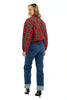 A woman stands smiling with her back to the camera, wearing Katydids Royal Stewart Plaid Holiday Quilted Jacket, blue cuffed jeans, and black heeled boots against a white background.