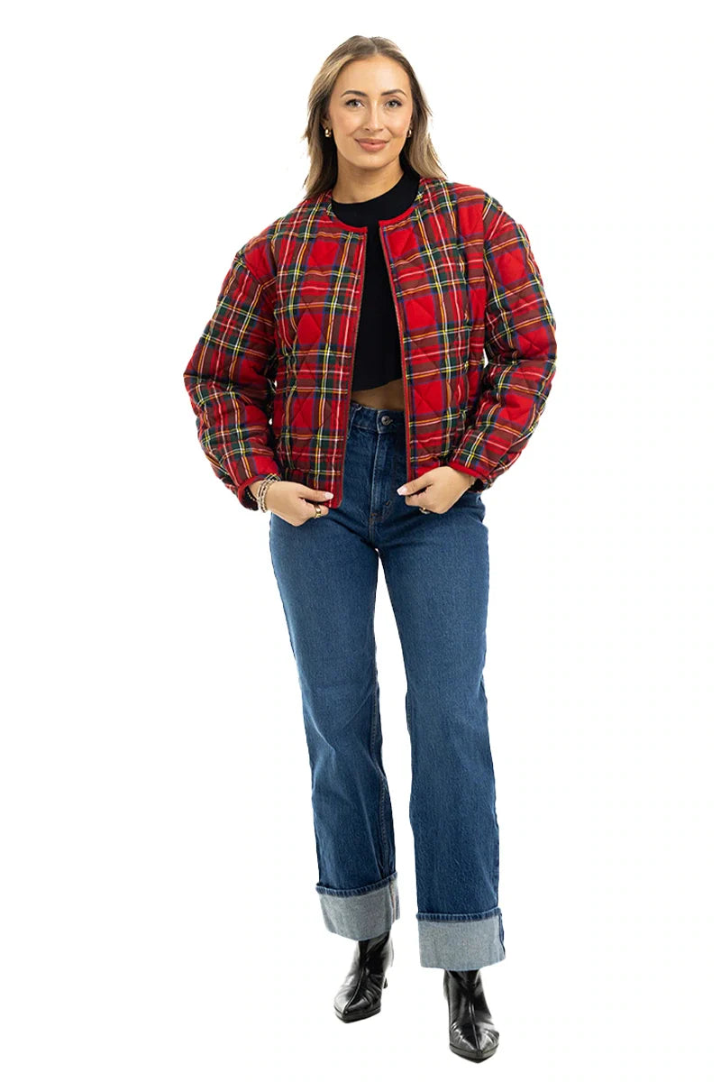 A woman stands smiling in a Katydid Royal Stewart Plaid Holiday Quilted Jacket over a black top, blue cuffed jeans, and black boots against a white background.