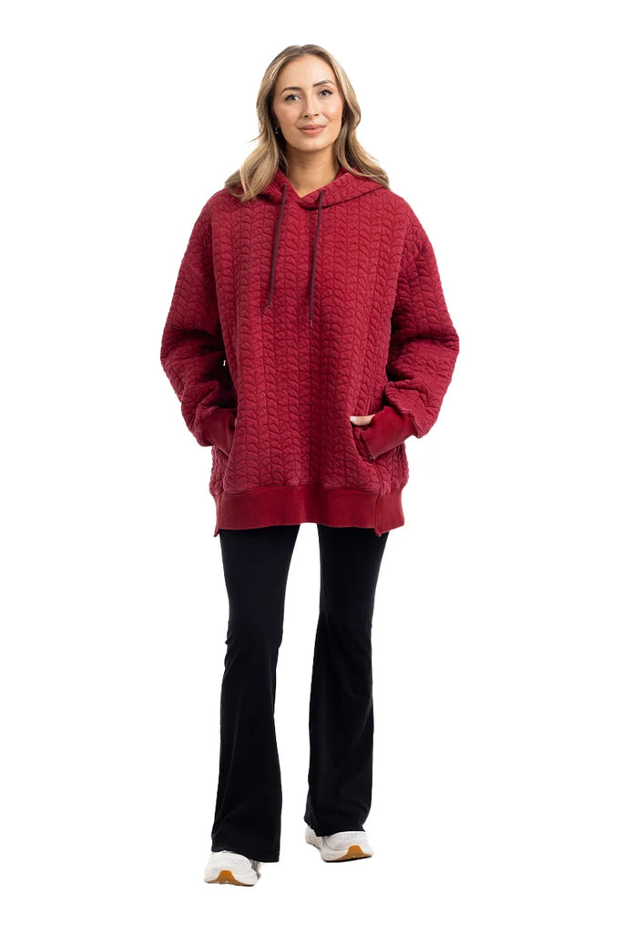 A woman smiles facing forward, hands in pockets, wearing the Katydid Red Heart Quilted Garment Washed Hoodie for Women, paired with black flared pants and white sneakers.