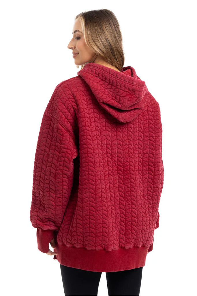 A woman with long, light brown hair wears the Katydid Red Heart Quilted Garment Washed Hoodie for Women and black leggings, facing away and glancing sideways against a plain white background.