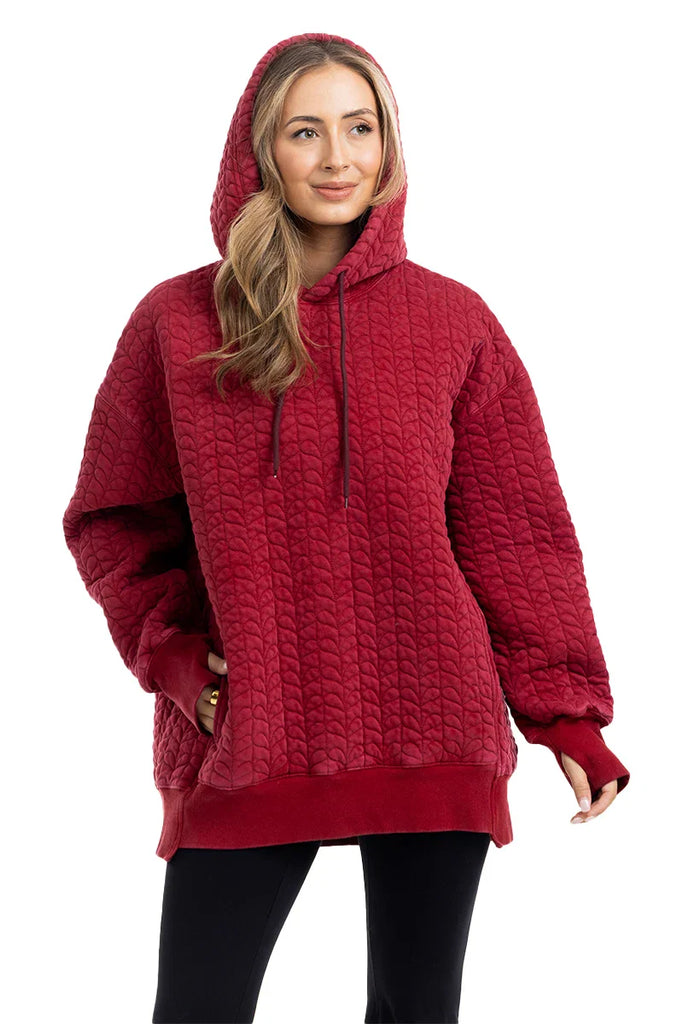 A woman models the Katydid Red Heart Quilted Garment Washed Hoodie for Women, featuring a hood, drawstrings, and front pocket. She smiles gently with one hand in her pocket, pairing the casual hoodie with black pants.