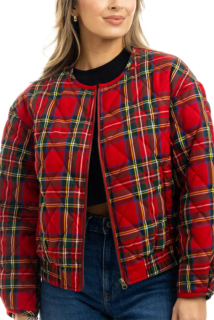A person with light brown hair, smiling slightly, wears the Katydid Royal Stewart Plaid Holiday Quilted Jacket over a black top and blue jeans. Shown from shoulders to waist.