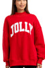 A woman smiles in front of a simple background, wearing the Katydid JOLLY Mockneck Sweatshirt—a bright red top featuring “JOLLY” in bold white letters—perfect for adding holiday cheer to her look.