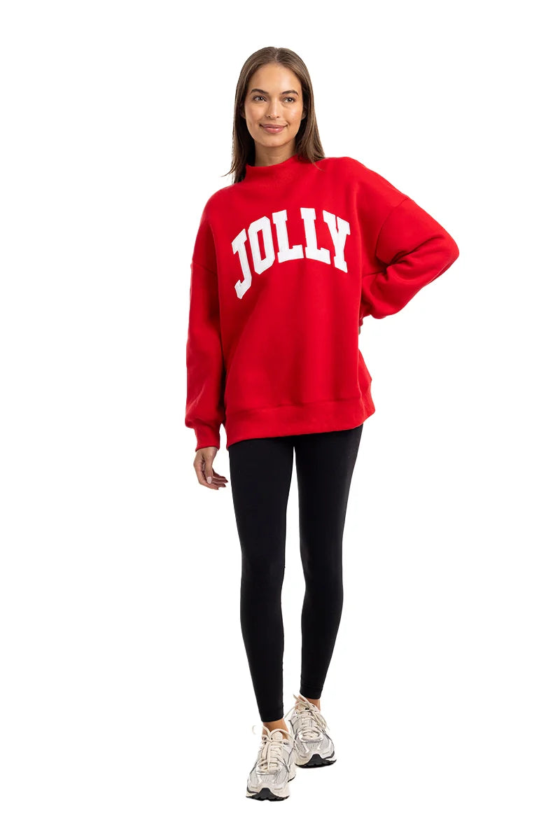 A woman smiles in the Katydid JOLLY Mockneck Sweatshirt—bright red with bold white letters—paired with black leggings and white sneakers, posed against a plain white background.