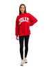A woman smiles in the Katydid JOLLY Mockneck Sweatshirt—bright red with bold white letters—paired with black leggings and white sneakers, posed against a plain white background.