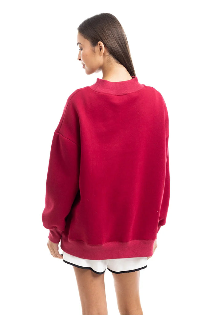 A woman faces away, wearing the Katydid Red Essential Solid Mockneck pullover sweatshirt paired with red and white shorts with black trim—a simple, stylish fall look set against a plain white background.