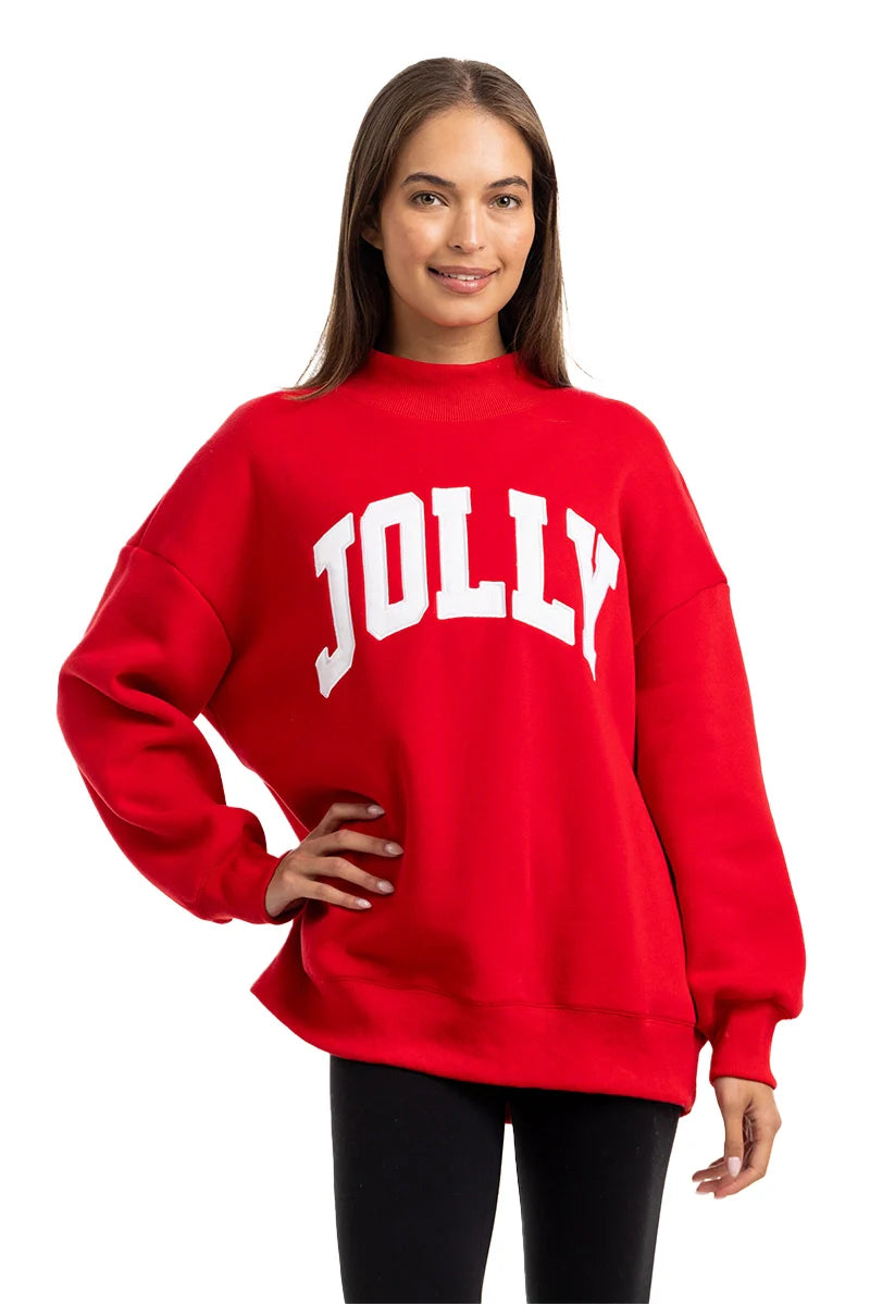 A person with long brown hair smiles while wearing the Katydid JOLLY Mockneck Sweatshirt, a bright red pullover with bold white letters on the front, paired with black pants and set against a plain white background.