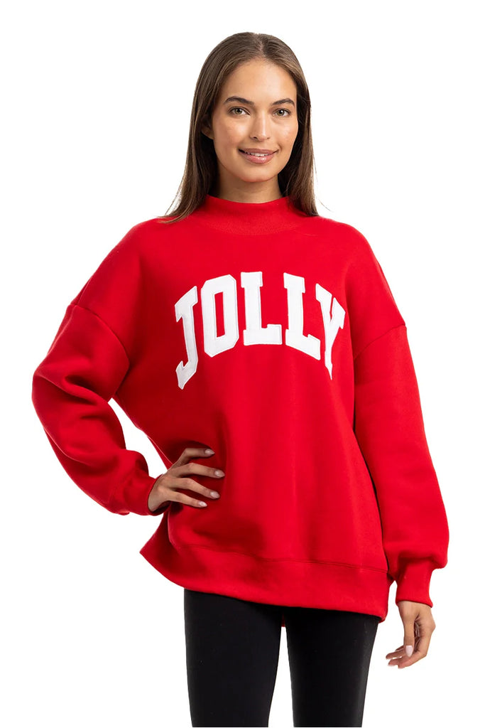 A person with long brown hair smiles while wearing the Katydid JOLLY Mockneck Sweatshirt, a bright red pullover with bold white letters on the front, paired with black pants and set against a plain white background.