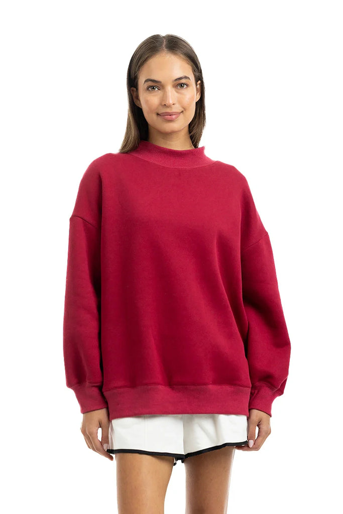 A woman with straight brown hair smiles gently while wearing the Katydid Red Essential Solid Mockneck pullover and white shorts, standing against a white background—a perfect cozy look for your fall wardrobe.