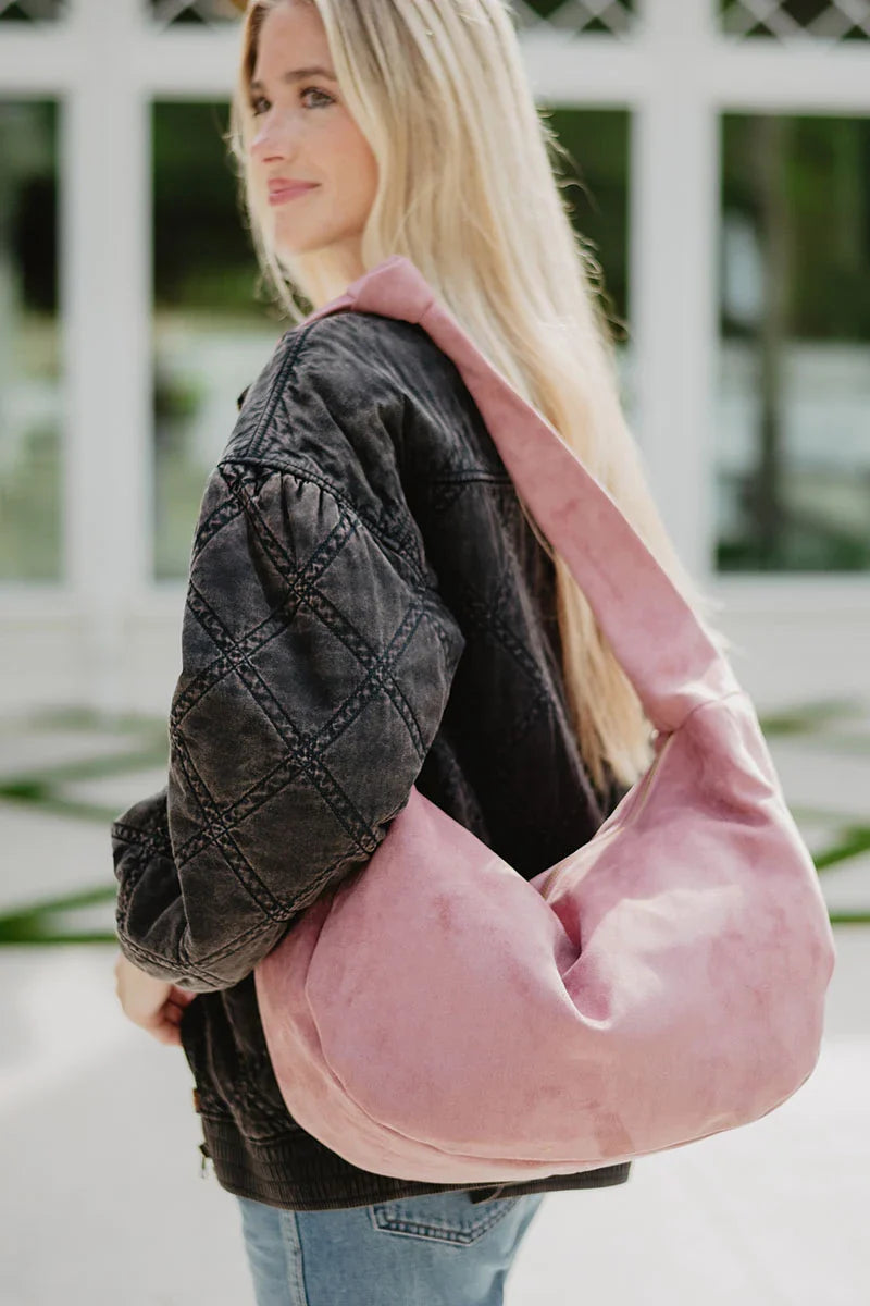 A woman with long blonde hair wears a black quilted jacket and blue jeans, carrying the Katydid Blush Suede Tote Bag. She stands outdoors in front of a white building with large windows.
