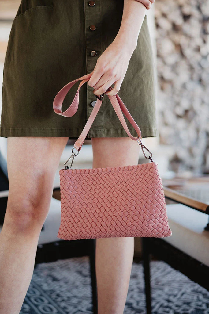 A person holds a Katydid Blush Woven Everywhere Crossbody Bag with an adjustable strap in their right hand, wearing an olive green buttoned dress. The blurred background features indoor elements and stacked firewood.