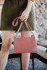 A person holds a Katydid Blush Woven Everywhere Crossbody Bag with an adjustable strap in their right hand, wearing an olive green buttoned dress. The blurred background features indoor elements and stacked firewood.